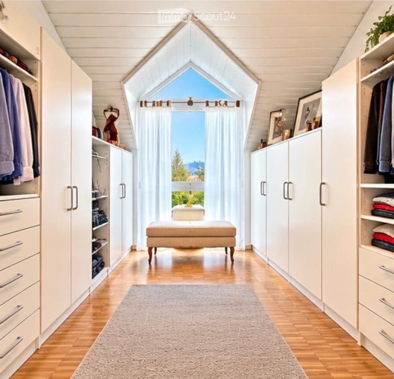 Bright, airy walk-in closet with wooden floors, white built-in wardrobes, a small bench, and a large skylight window offering a scenic view.