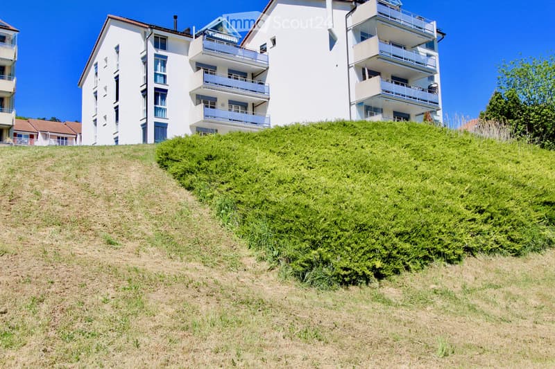 Multi-story residential building with white exterior, multiple balconies, and a sloped green hillside in front.