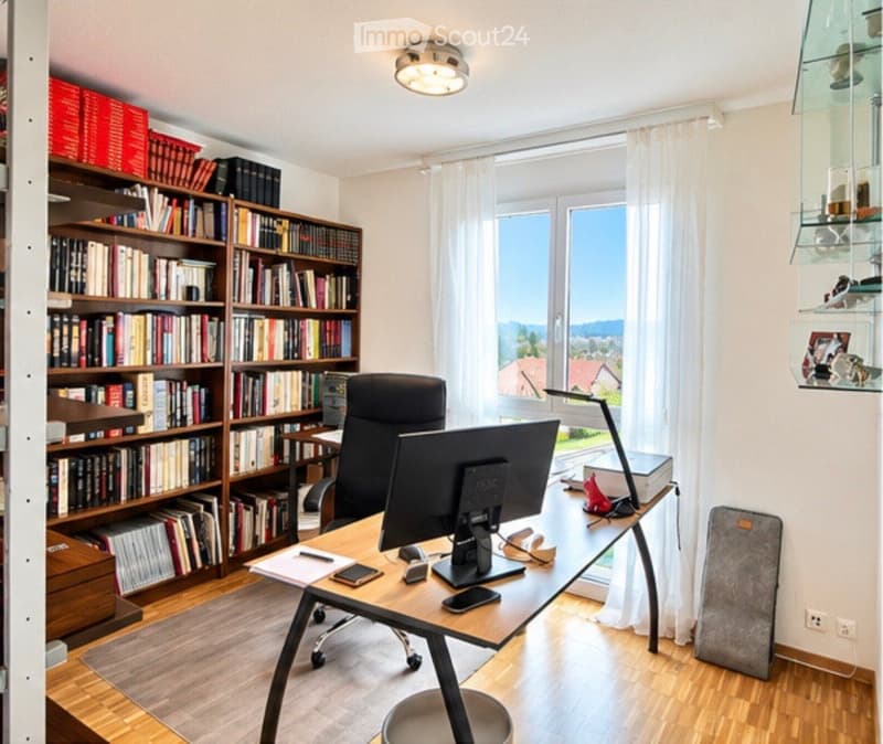 A well-organized home office with a wooden desk, ergonomic chair, computer monitor, bookshelf filled with books, and a large window with white curtains offering a scenic view.