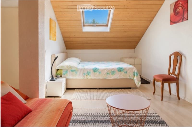 Cozy attic bedroom with a slanted wooden ceiling, skylight window, bed with floral bedding, nightstand with lamp, small sofa, wooden chair, and round table.