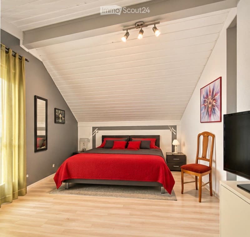 Modern bedroom with wooden flooring, slanted white ceiling, red bedding, grey walls, and a large window with yellow curtains.