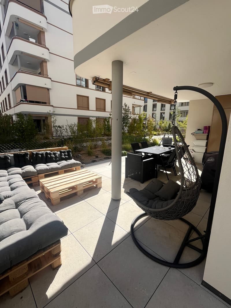Balcony with a grey sectional sofa, wooden coffee table, wicker hanging chair, and a small dining area with a table and chairs.