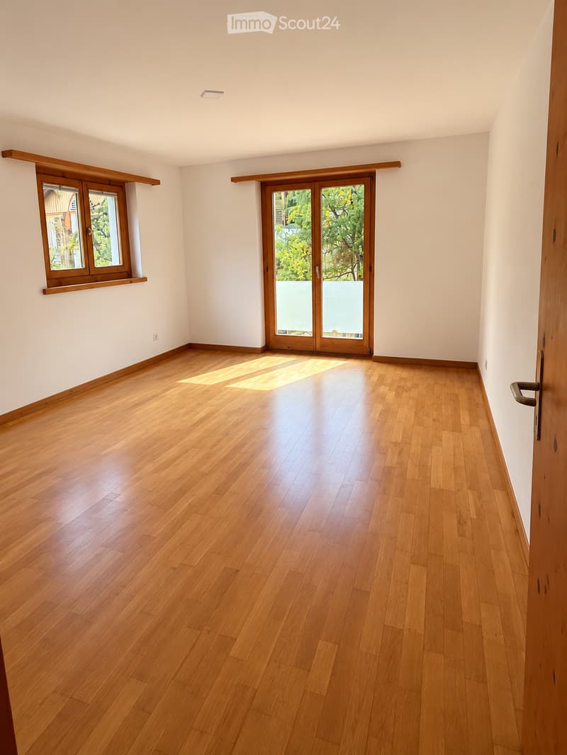 Empty room with wooden flooring, white walls, and large windows with wooden frames. Sunlight streams in through the windows, illuminating the space.