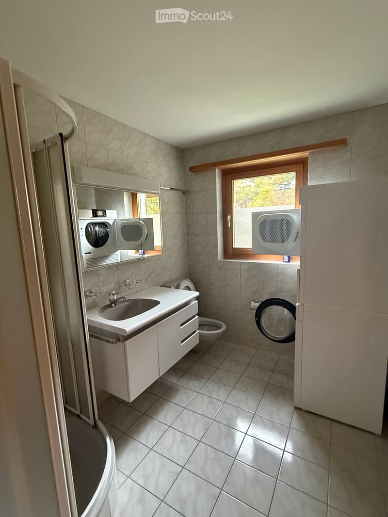 Modern bathroom with tiled walls and floor, white sink with cabinet, toilet, mirror, and a washing machine and dryer unit.
