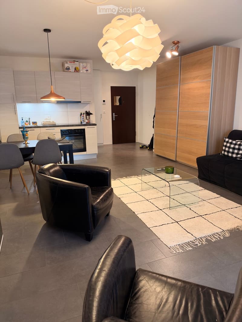 Modern open-plan living and dining area with a black leather sofa, glass coffee table, dining table with chairs, and a kitchen area with white cabinets and stainless steel appliances.