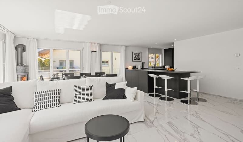 Modern living and dining area with white sofa, black coffee table, kitchen island with bar stools, fireplace, and large windows with white curtains.