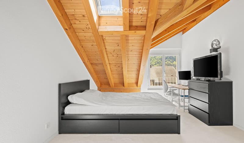 Modern bedroom with a slanted wooden ceiling, large window offering a view, a black bed, a nightstand, a desk with a chair, a laptop, and a television on a stand.