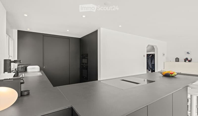 Modern kitchen with grey countertops, white walls, and built-in appliances. Features a sink, faucet, oven, fridge, and a fruit bowl on the counter.