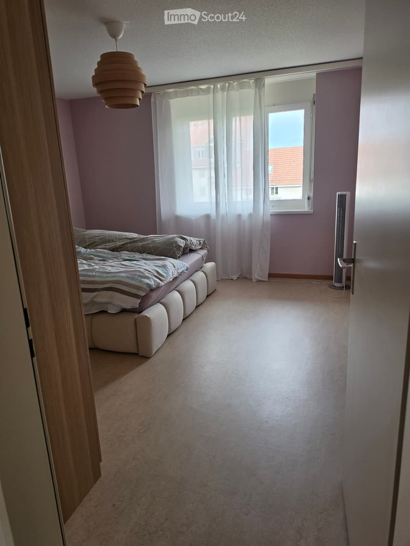 Bedroom with a bed, fan, and large window with curtains. Light purple walls and carpeted floor.