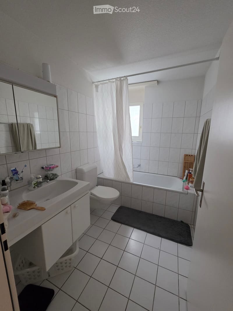 Bathroom with white tiles, a sink, toilet, bathtub, mirror, and a shower curtain. Includes a towel, bathroom mat, and personal care items on the sink counter.