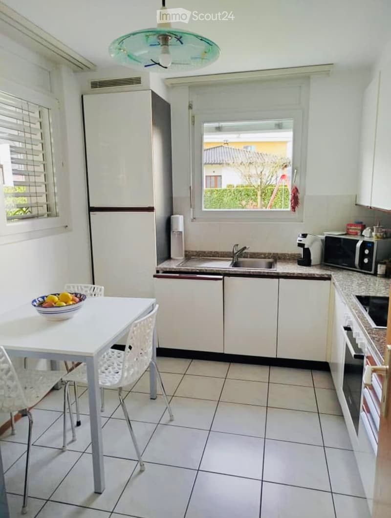 Bright kitchen with white cabinets, tiled floor, and a dining area featuring a white table and chairs. Appliances include a refrigerator, microwave, and coffee maker.