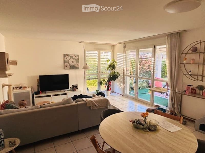 Bright living room with large windows and a view of the garden. Features a grey sofa, coffee table, TV, dining table with chairs, and decorative plants.