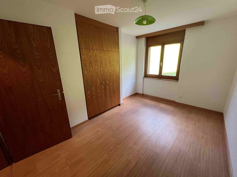 Empty room with wooden flooring, white walls, and a window letting in natural light. There are two wooden doors and a green hanging light fixture.