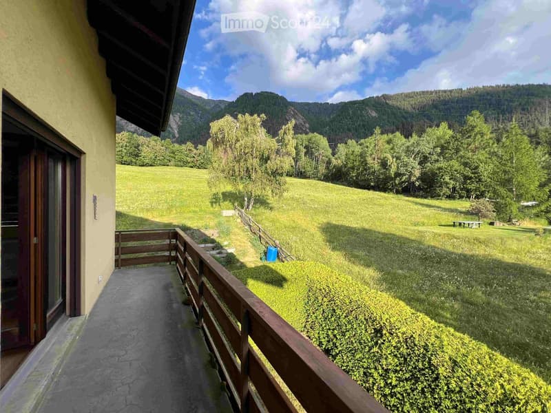 Balcony with wooden railing overlooking a green field and forested hills under a partly cloudy sky.