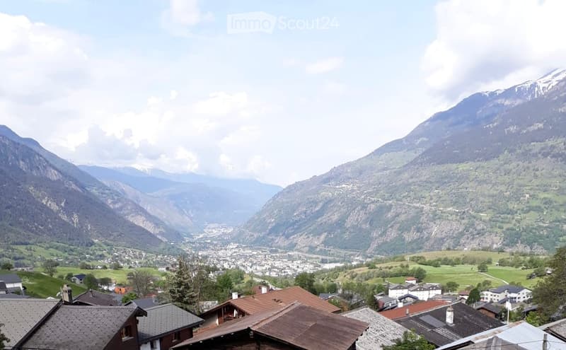 Panoramic view of a village nestled in a valley surrounded by mountains, with visible houses, green fields, and a partly cloudy sky.