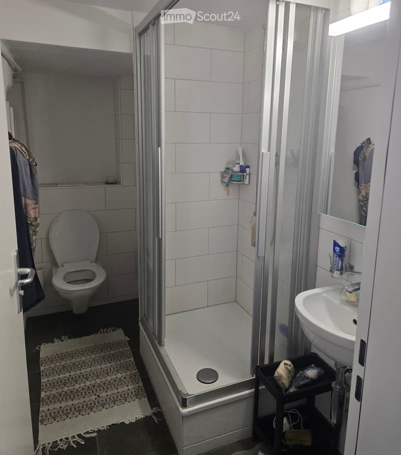A small bathroom with white tiles, a toilet, a sink, a shower, a rug, and a towel hanging on the wall.