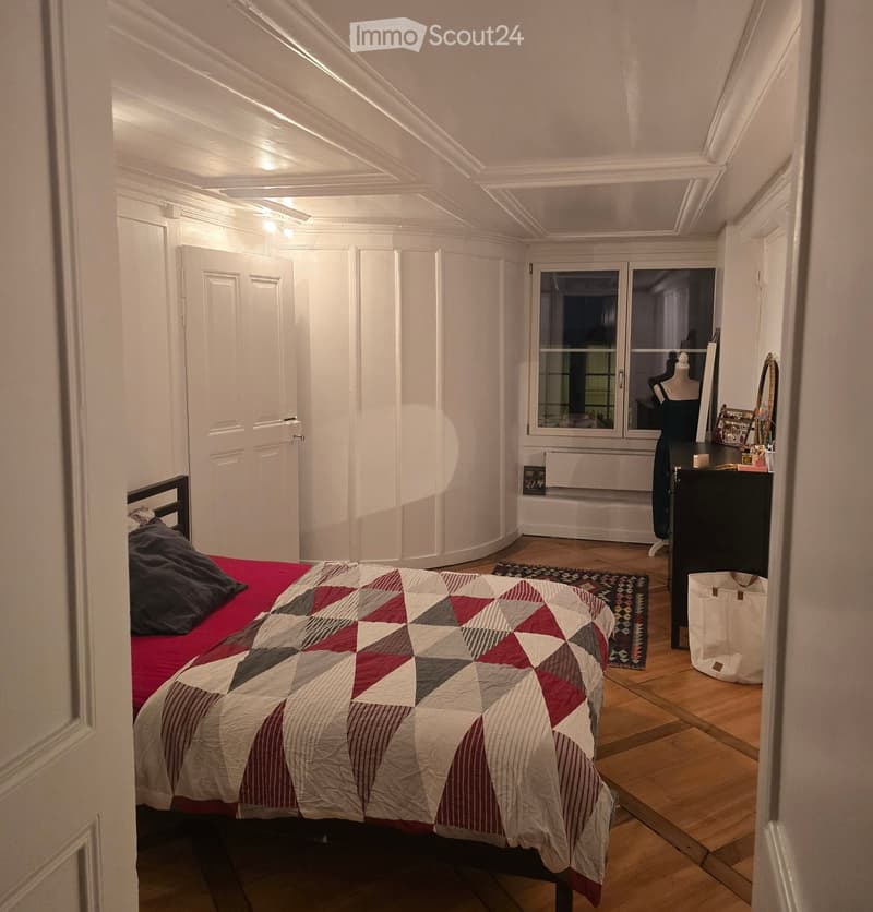 Cozy bedroom with wooden flooring, a bed with a red and white quilt, a nightstand with a mirror, and a small table with a lamp.