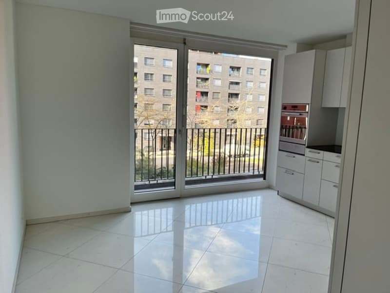 Bright, unfurnished room with large sliding glass doors opening to a balcony, white tiled floor, modern white kitchen cabinets visible, view of a city building.