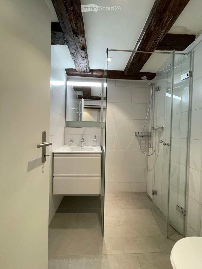 Modern bathroom with white tiles, exposed wooden beams, a glass-enclosed shower, a sink with a mirror, and a cabinet.