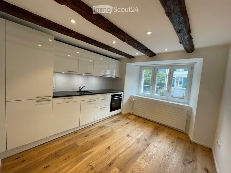 Modern kitchen with light-colored cabinets, a black countertop, and wooden flooring. Features include a sink, oven, and a window with a radiator below.