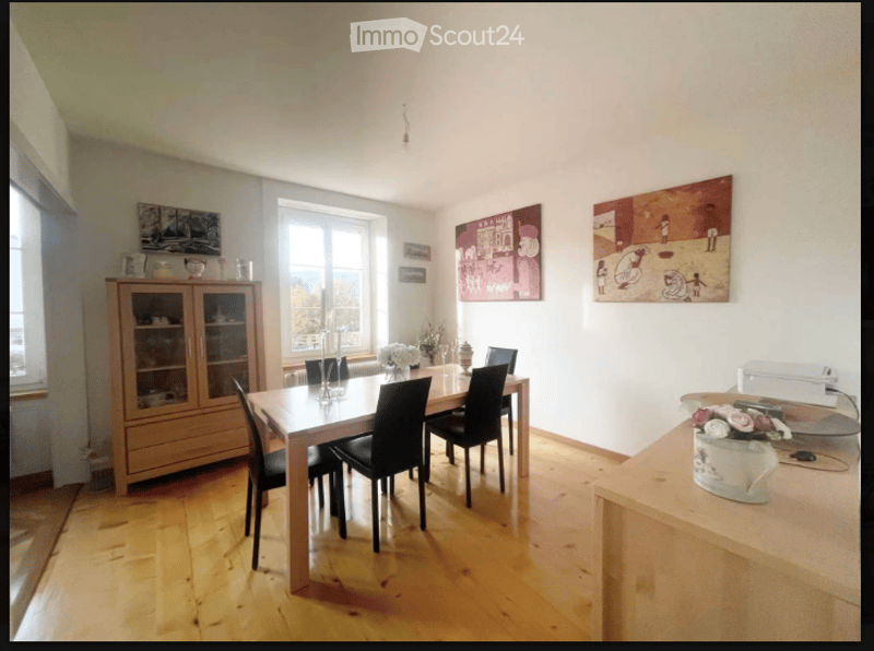 Bright dining room with wooden flooring, large windows offering a scenic view, a wooden dining table surrounded by black chairs, a wooden cabinet with glass doors, and decorative vases on the table and counter.