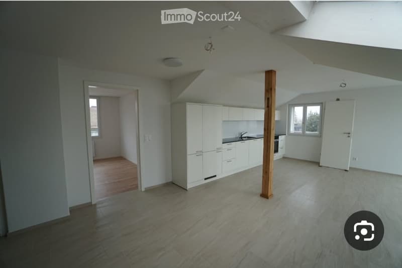 Empty room with white walls, wooden floor, and white kitchen cabinets visible in the background.