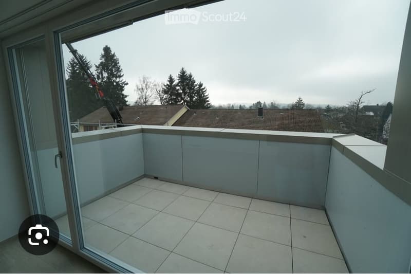 Balcony with tiled floor, white walls, and a view of trees and rooftops.