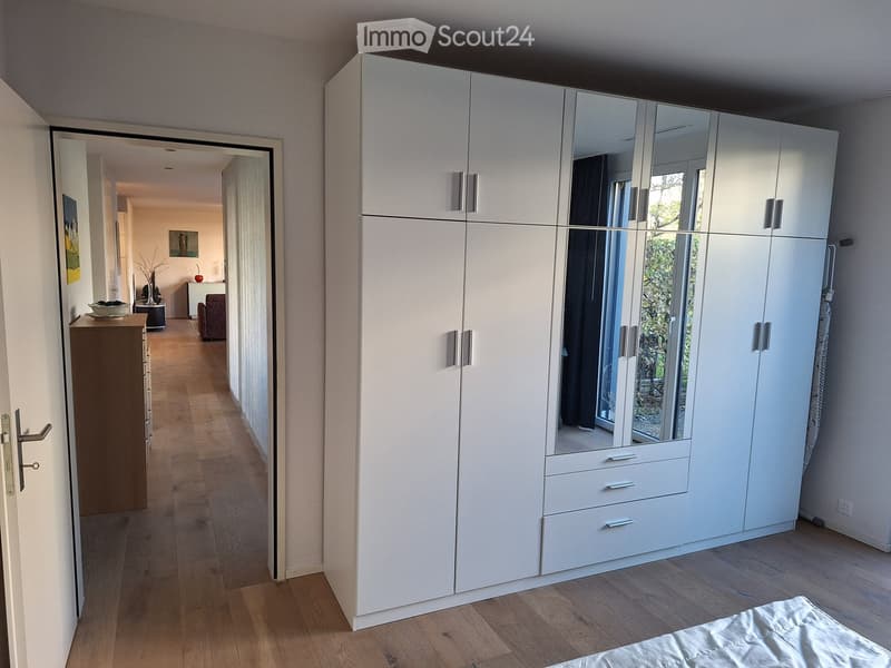Spacious bedroom with a large white wardrobe with mirrored doors, a neatly made bed, and a hallway leading to another room with a small table and chair.