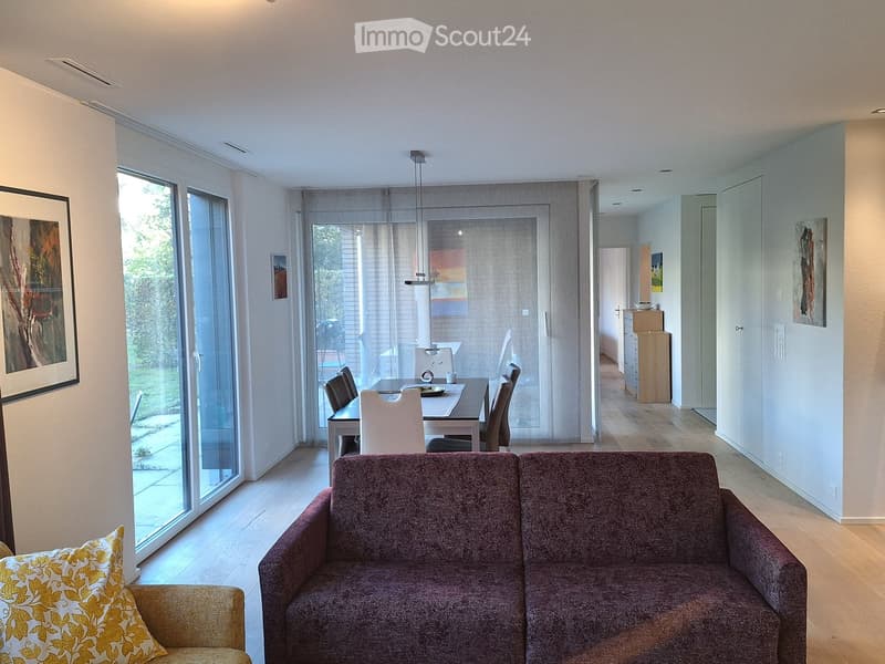 Modern living and dining area with a sofa, dining table, chairs, and a small cabinet. Large windows and sliding glass doors lead to an outdoor space.