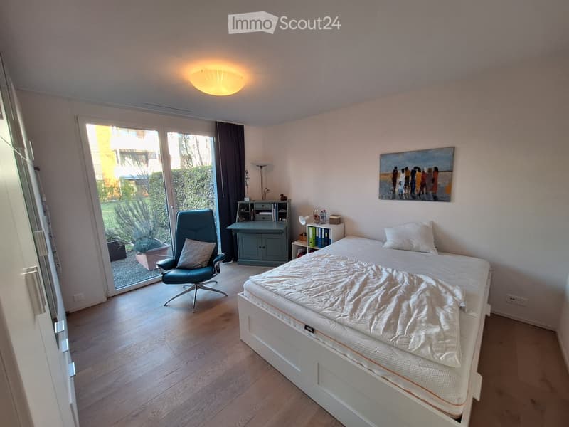 Bright bedroom with wooden flooring, large bed, armchair, small cabinet, and artwork on the wall. Large window with view to the garden.