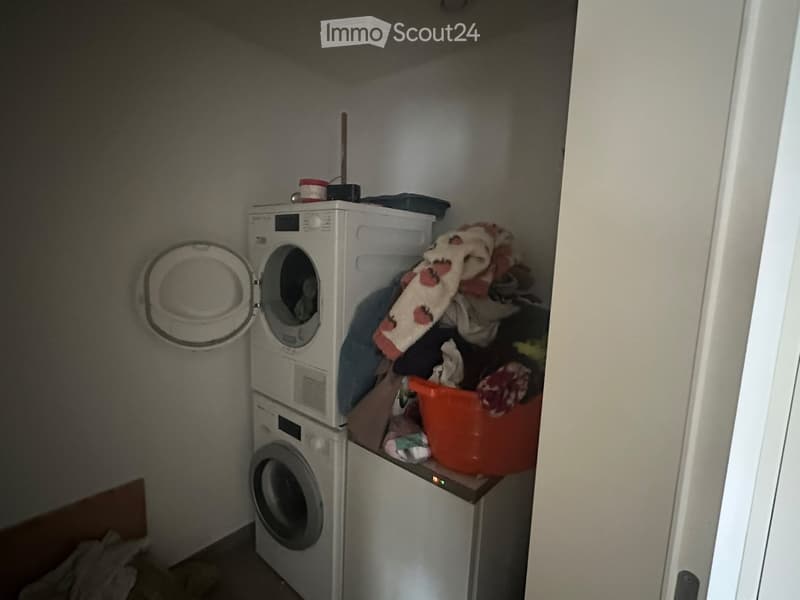 A small laundry area with a stacked washing machine and dryer, an orange bucket, and some folded clothes on top of the dryer.