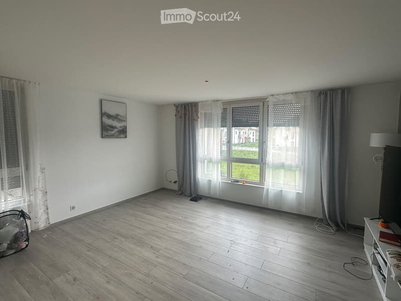 Empty room with wooden flooring, large windows with sheer curtains, and a few small items like a picture frame and a small basket on the floor.