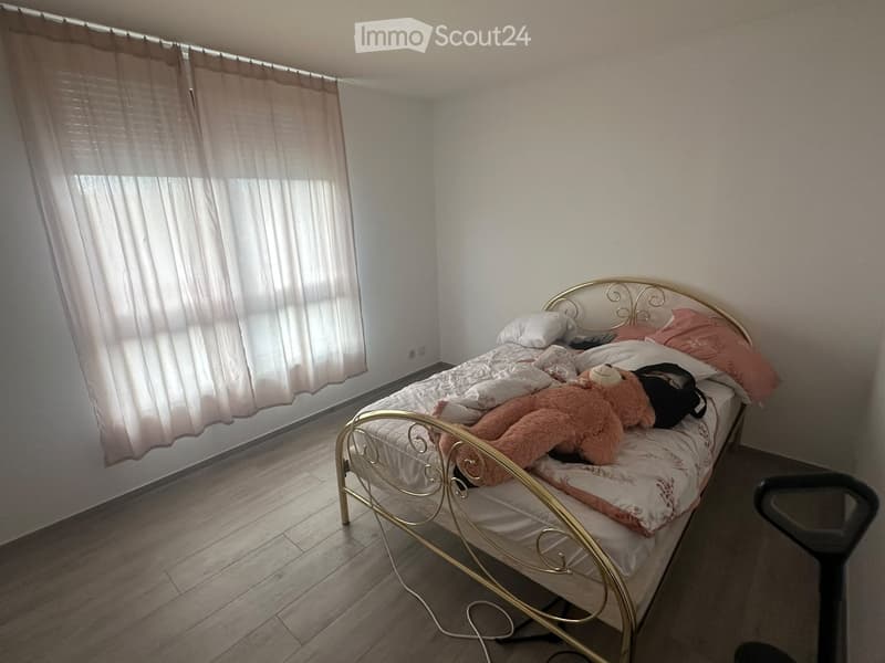 A small bedroom with a single bed, pillows, a teddy bear, and a backpack on the bed. The room has wooden flooring and white walls with a window covered by light-colored curtains.