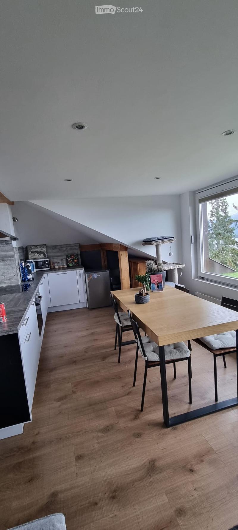 Open-plan kitchen and dining area with wooden flooring, white cabinets, and modern appliances. A dining table with chairs is set near a large window offering a view of greenery.