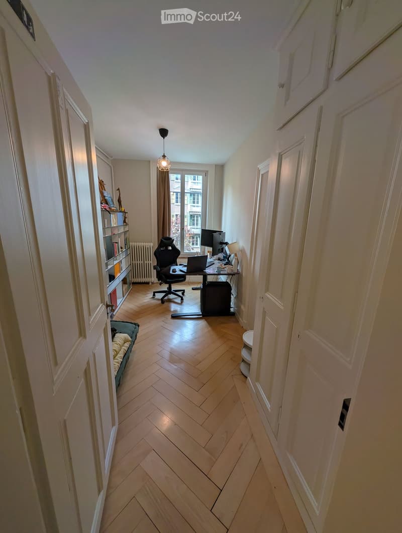 A furnished room with wooden herringbone flooring, a desk with a laptop and screen, a chair, a shelf with books and decorative items, a radiator, and a pet bed.