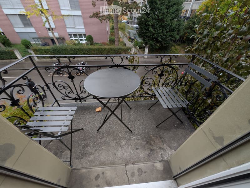 Balcony with a round metal table and two metal chairs, overlooking a street with buildings and trees.