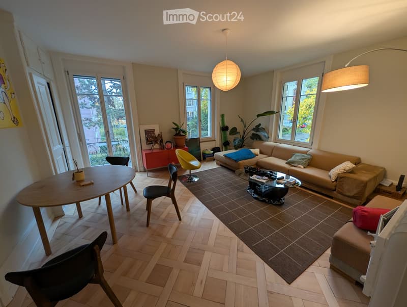Bright living room with wooden flooring, large windows, a beige sofa, coffee table, and a round wooden dining table with chairs.