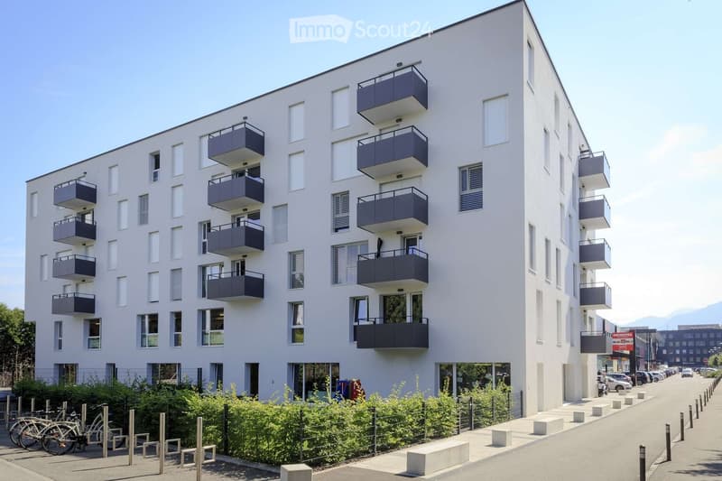 Modern multi-story apartment building with white facade and dark grey balconies, located on a street with parked cars and bicycles.