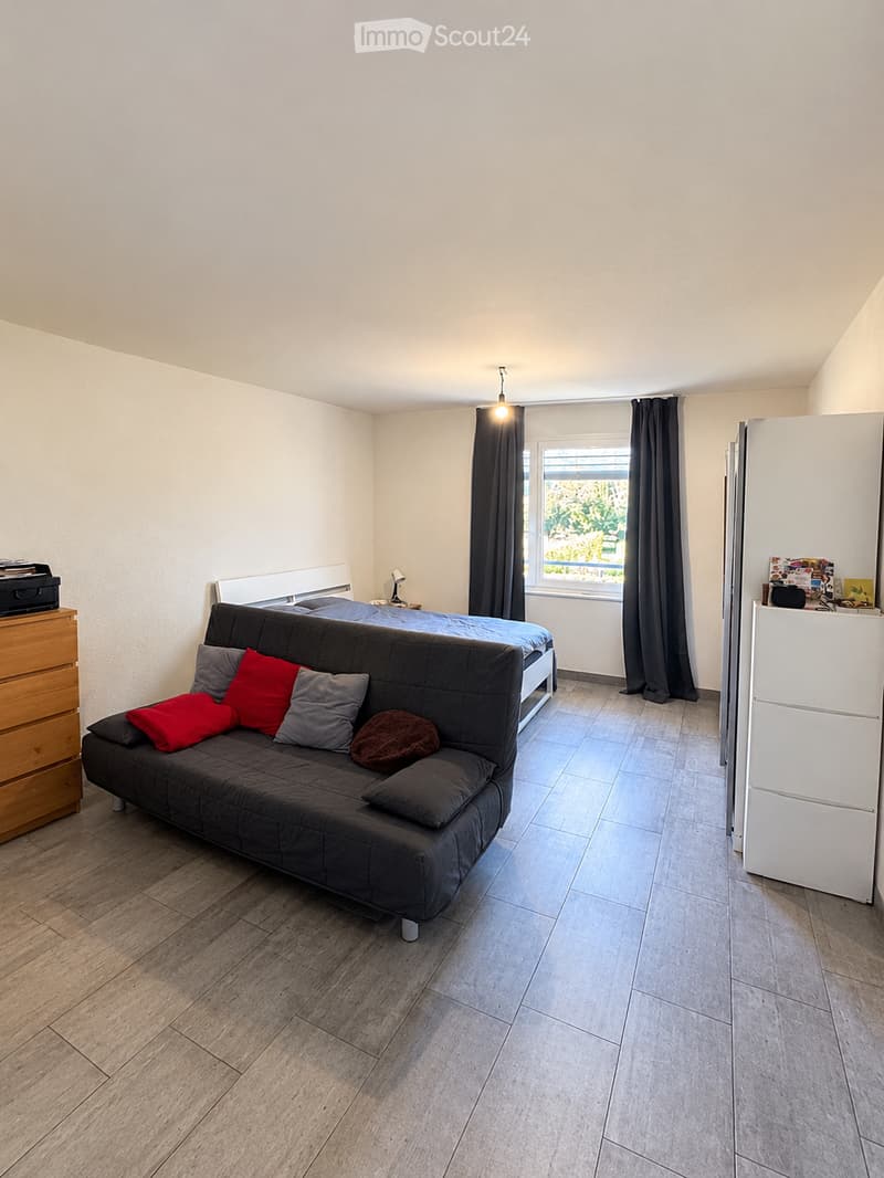 A modern, compact bedroom with a sofa, bed, nightstand, and a refrigerator. The room features wooden flooring, white walls, and a large window with dark curtains.