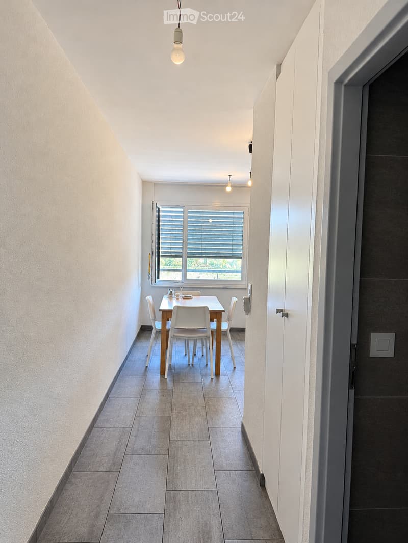 A modern, minimalist dining area with a wooden dining table and white chairs, situated near a large window with blinds, allowing natural light to fill the space.