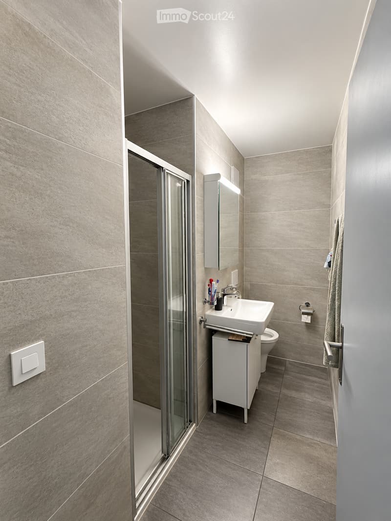 Modern bathroom with a white sink, mirror, toilet, and glass-enclosed shower. Beige tiles on walls and floor, towel hanging on the side.