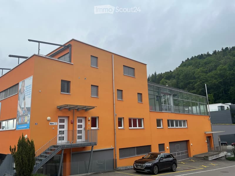 Modern two-story building with orange facade, multiple windows, and a balcony. Features a staircase leading to an upper level and a parked car in front.