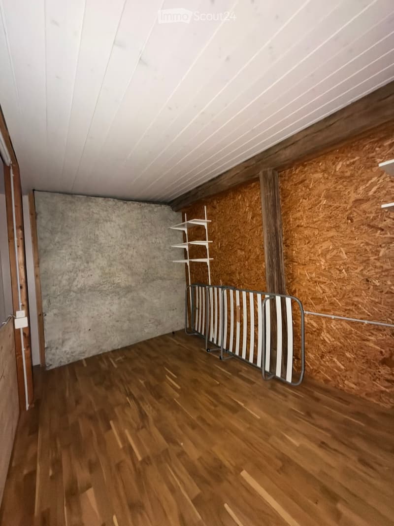 Empty room with wooden flooring, exposed wooden walls, and a slanted white ceiling. A metal gate and a white shelving unit are present.