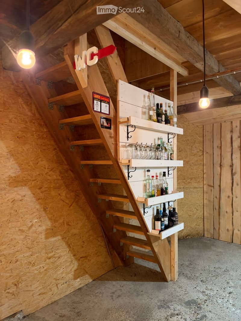 A wooden staircase leads to a small bar area with shelves holding various bottles and glasses, illuminated by hanging lights.