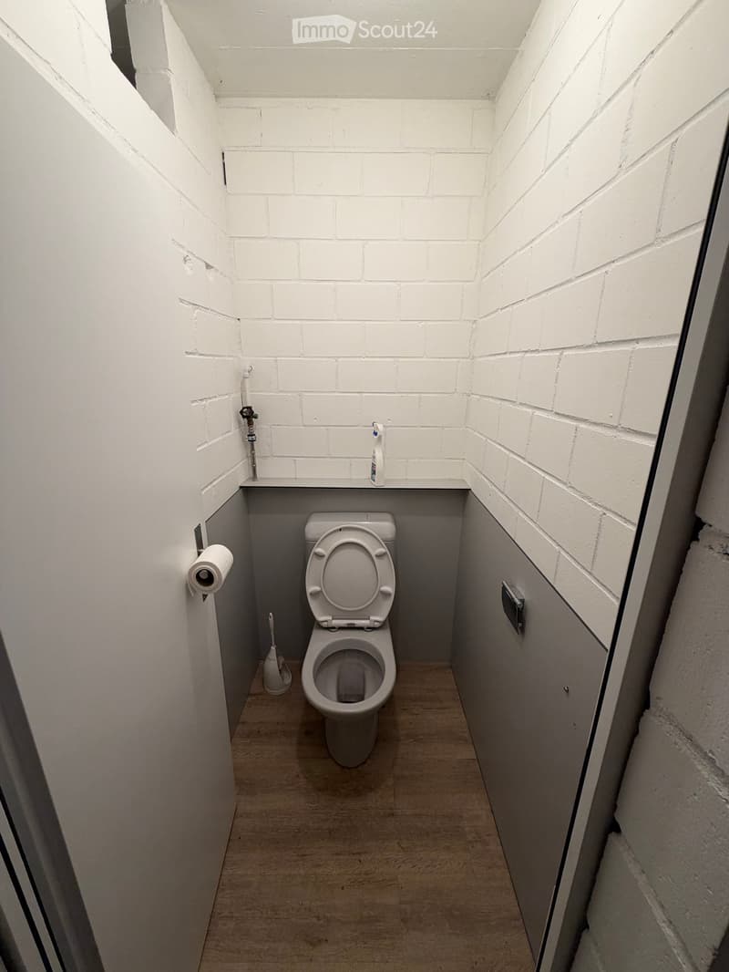Small bathroom with white brick walls, a toilet, toilet paper holder, and a toilet brush. The floor is light-colored wood.