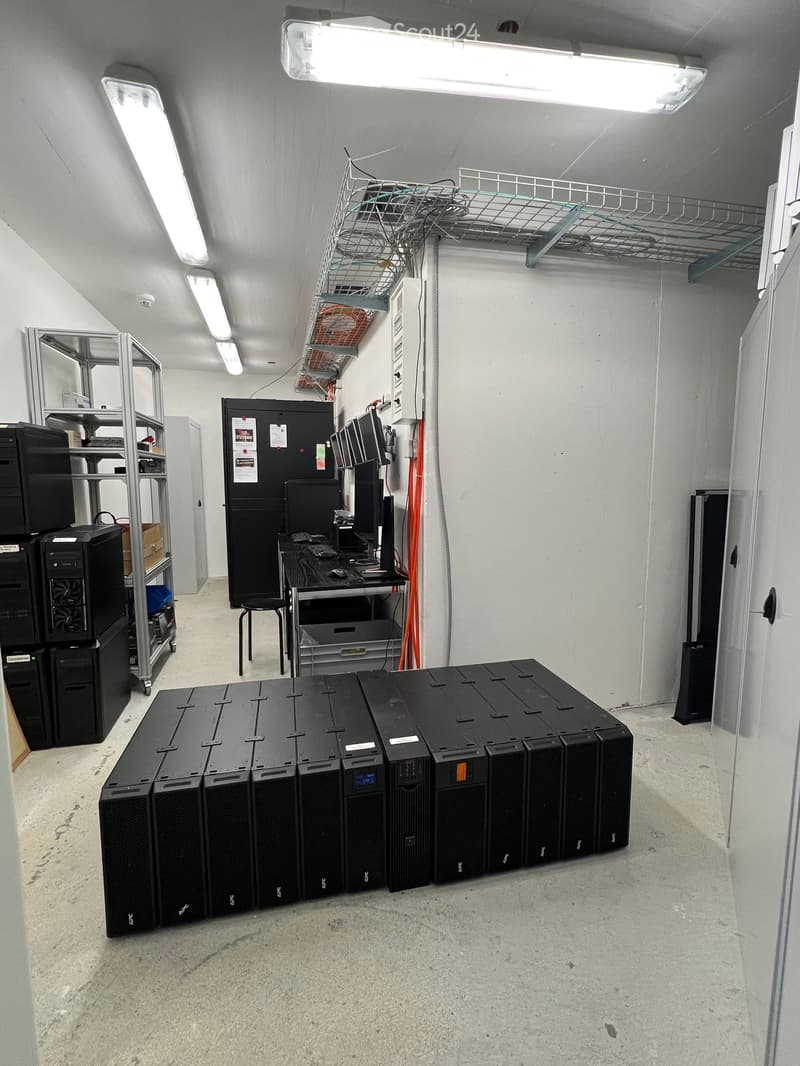 A server room with multiple server racks, a table with a computer setup, and a chair. The room has concrete floors and white walls with fluorescent lighting.