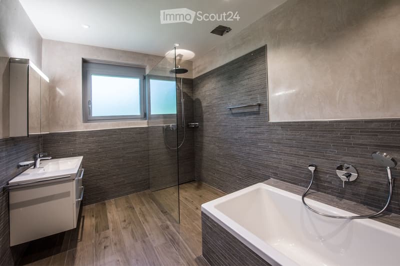 Modern bathroom with dark grey tiles, a bathtub, a sink, a shower, and a large window providing natural light.