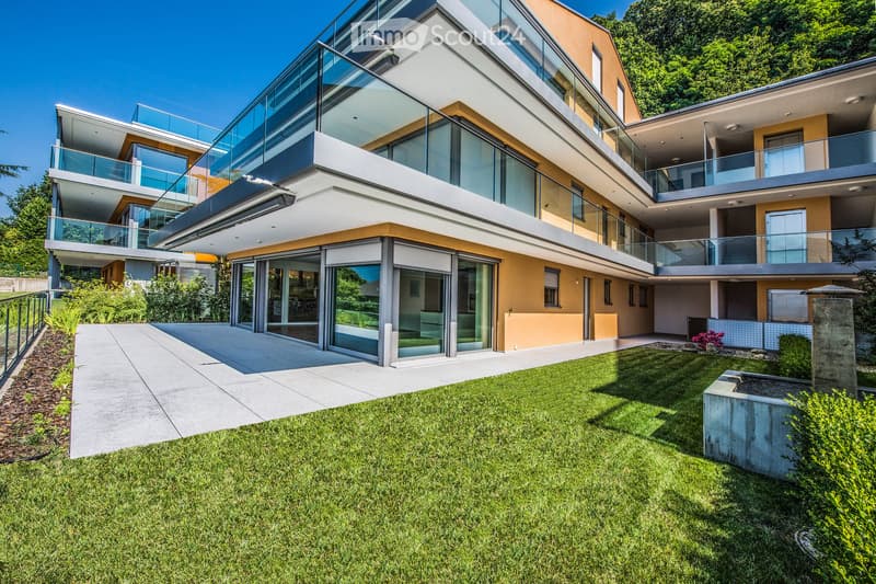 Modern two-story house with large glass windows and balconies, surrounded by a well-maintained lawn and small garden area.