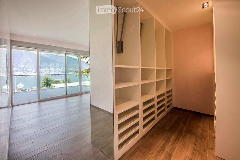 An unfurnished room with wooden flooring, large glass sliding doors leading to a balcony with a scenic mountain and lake view, and built-in white shelving units.