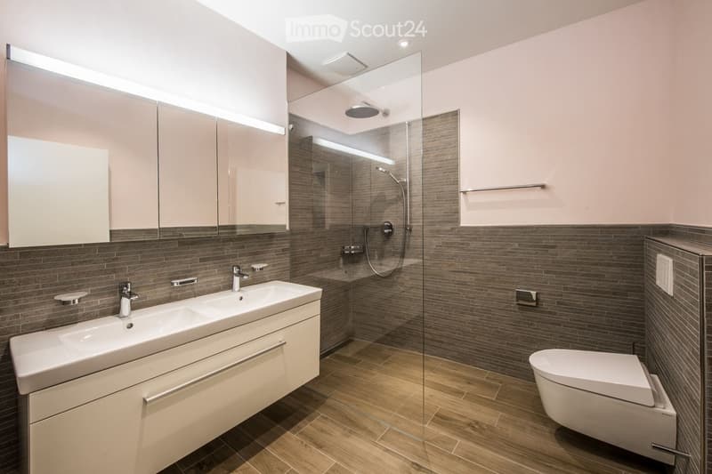 Modern bathroom with double sink, large mirror, glass-enclosed shower, and toilet. Features wooden flooring and grey tiled walls.